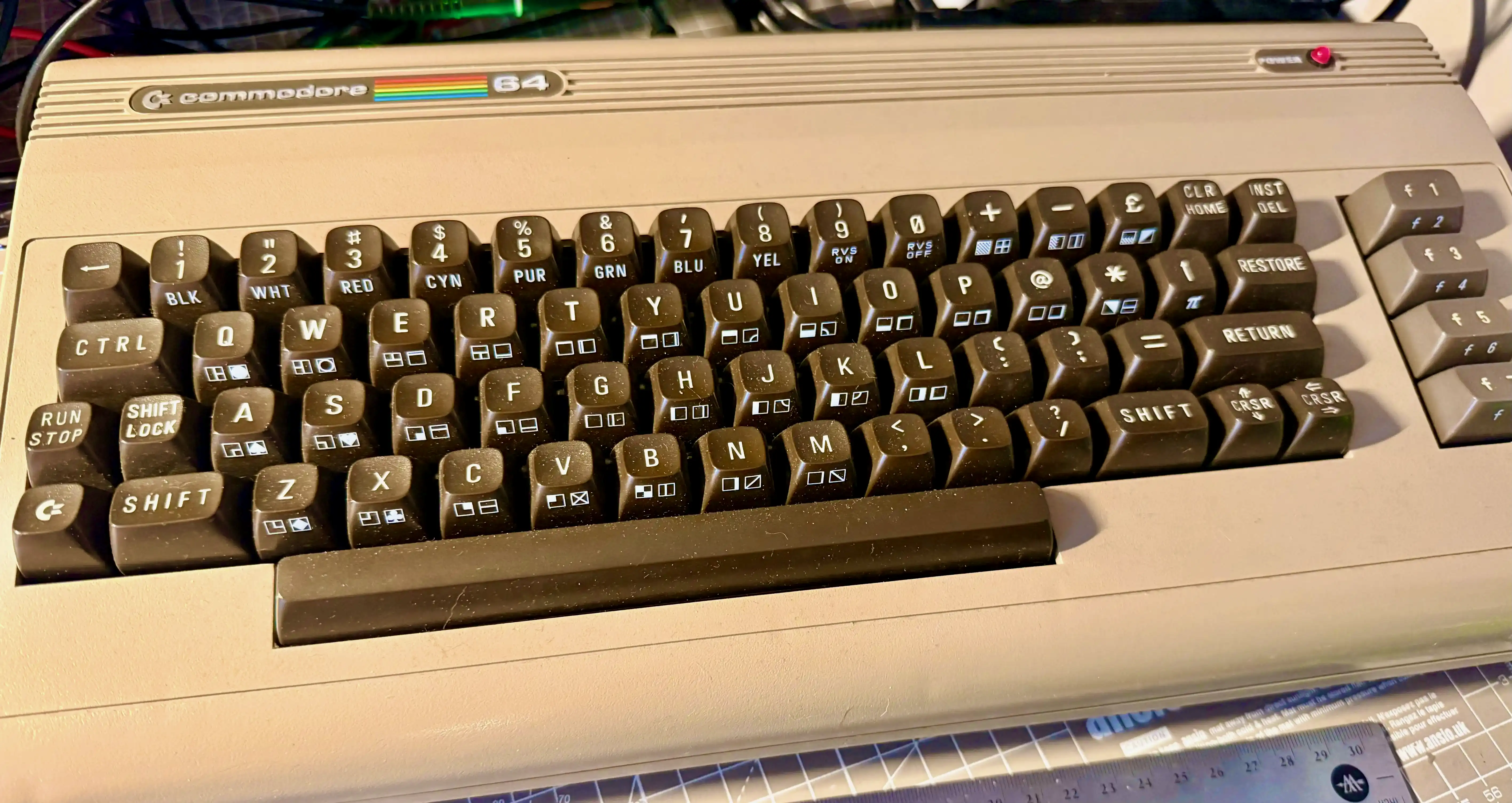 A well-used 1984 Commodore 64 breadbin with dusty keys, sitting on a cutting mat next to a ruler.